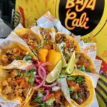 Baja Cali meat tacos set on a plate in a circle with limes and peppers in the center and the Baja Cali logo behind them