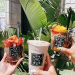 hands holding up three Acai Bar drinks with fruits and smoothies against a background of palm leaves