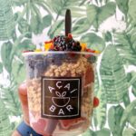a hand wearing a watch with a blue band holding up an Acai Bar fruit parfait with granola and blackberries with a wallpaper background of tropical leaves